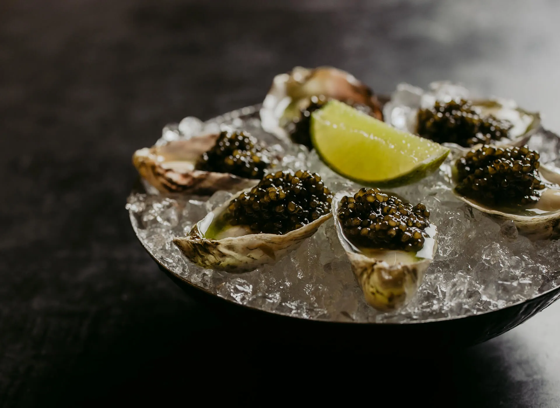 A close up shot of oysters with a spoon of caviar inside each, in a bowl of ice and garnished with a fresh lime.