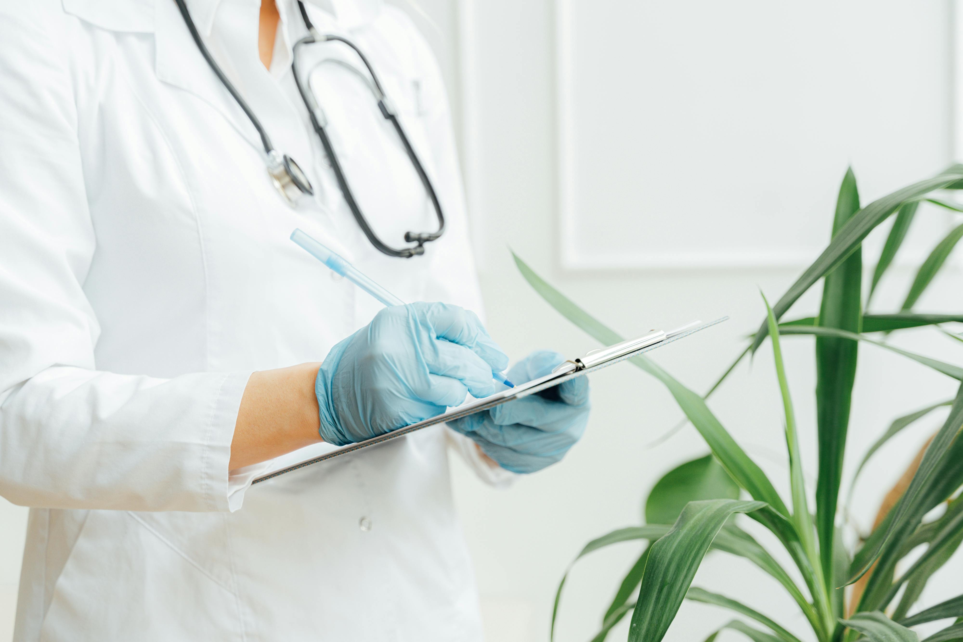 Medical professional wearing blue gloves and a white coat writing on a clipboard with a stethoscope around their neck.