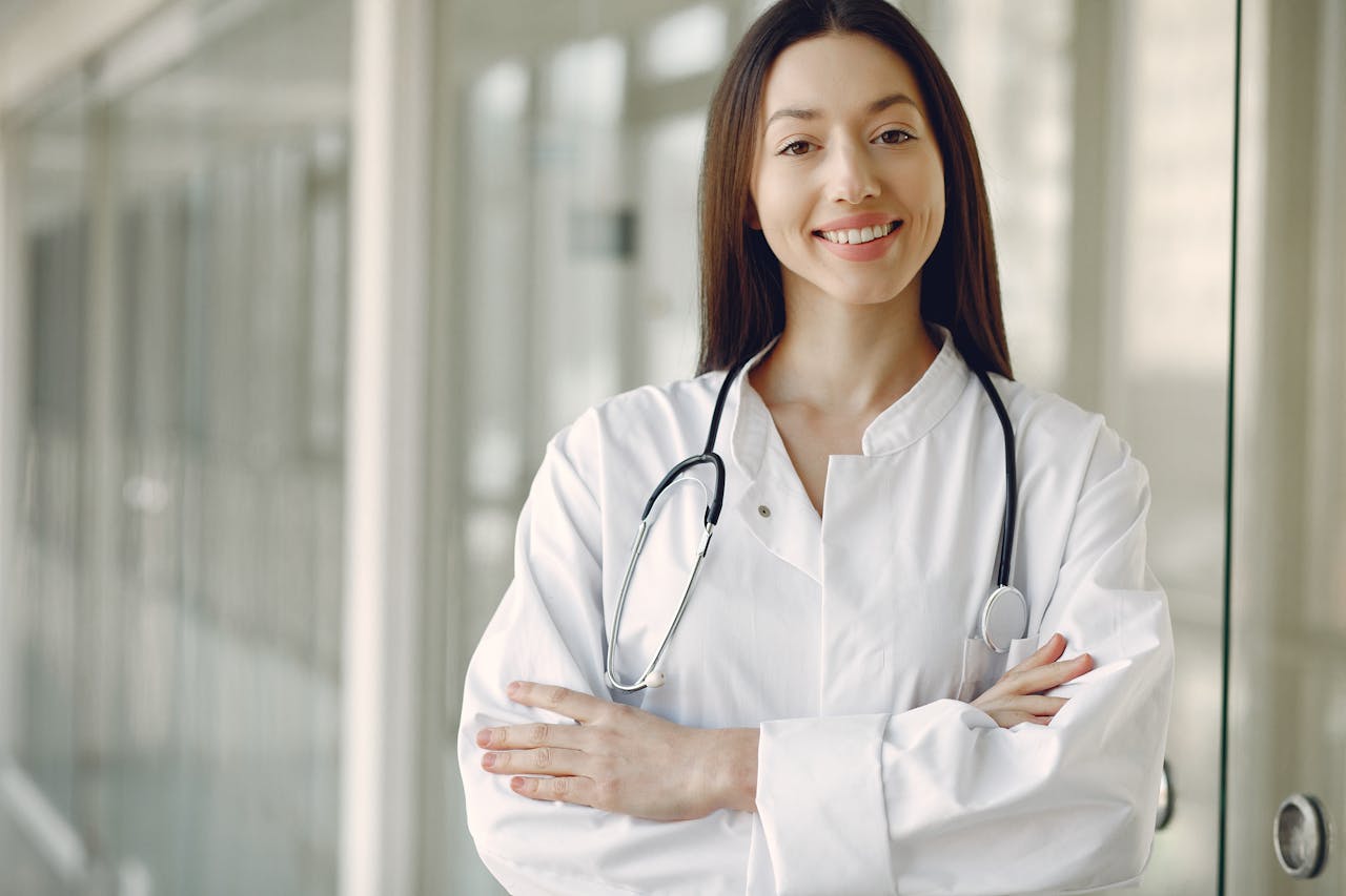 Smiling female doctor in white coat with stethoscope around neck standing with arms crossed.
