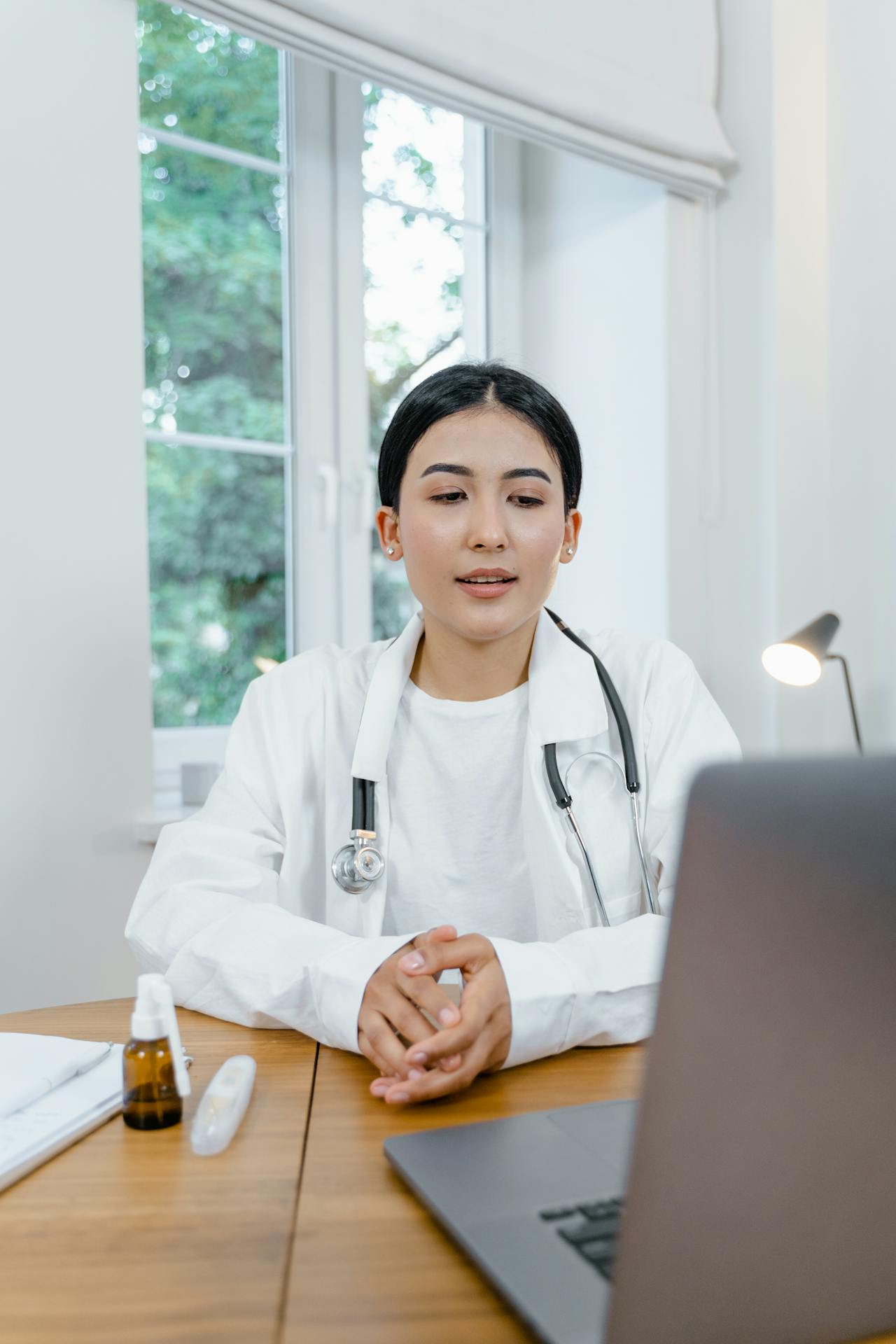 Female doctor wearing a white coat and stethoscope sits at a desk, looking at a laptop during a video consultation.