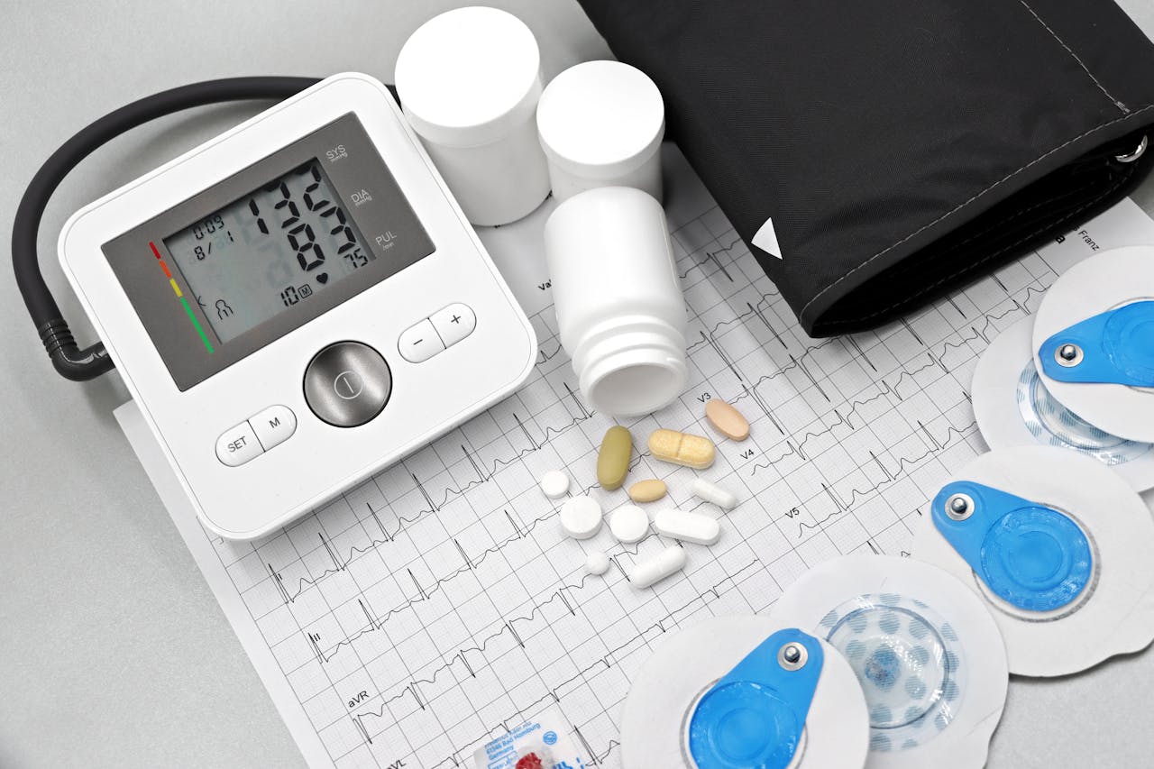 Digital blood pressure monitor displaying 132/83 with 75 pulse, surrounded by medicine bottles, pills, ECG paper, and heart electrode pads.