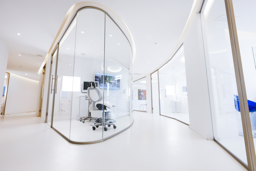 Modern, bright medical clinic hallway with curved glass-walled rooms and white floors.