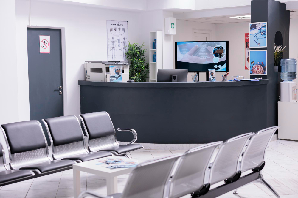 Modern clinic waiting area with metal chairs, a reception desk, a computer monitor, and medical posters on white walls.