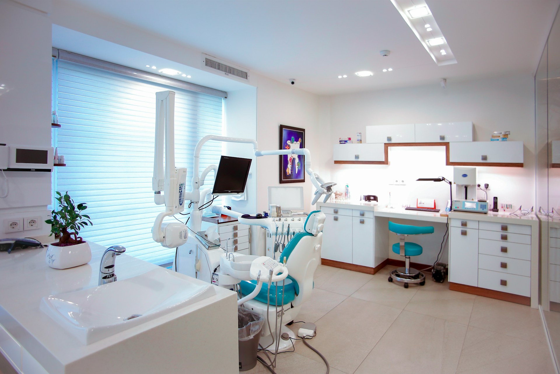 Bright modern dental clinic room with dental chair, equipment, a computer monitor, sink, and white cabinets.