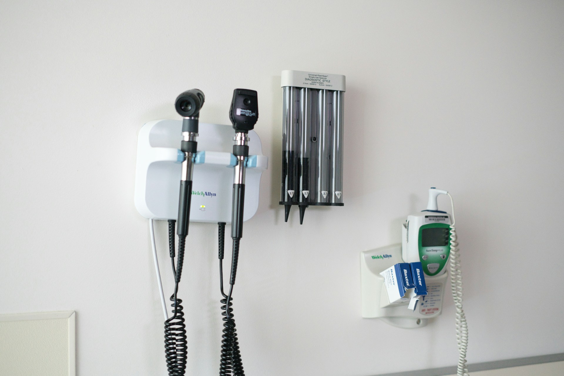 Wall-mounted medical diagnostic tools including an otoscope, ophthalmoscope, disposable specula holder, and an electronic thermometer in a clinical setting.