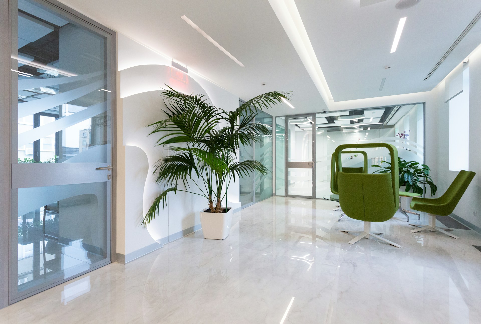 Modern office waiting area with green swivel chairs, glass doors, potted plants, and white marble flooring.