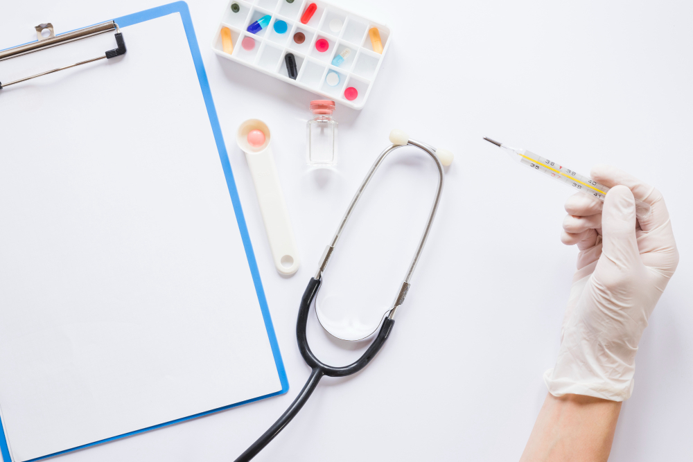 Gloved hand holding a mercury thermometer beside a stethoscope, medication blister pack, pill scoop, vial, and blank clipboard on a white surface.