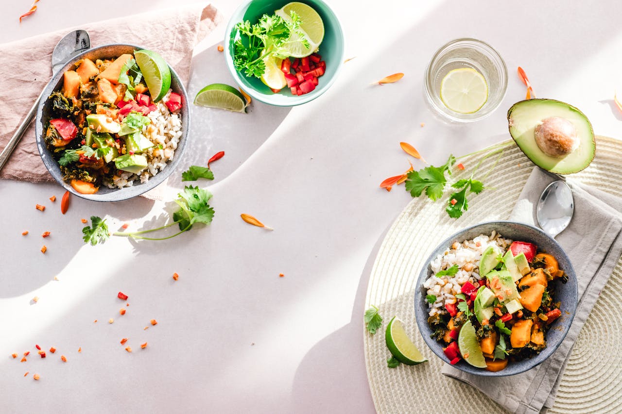 Two bowls of colorful vegetable and rice salad garnished with avocado, lime wedges, and cilantro on a light surface with a glass of lemon water and half an avocado.