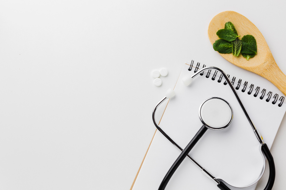 A stethoscope, a wooden spoon with green mint leaves, three white tablets, and a blank spiral notebook on a white surface.