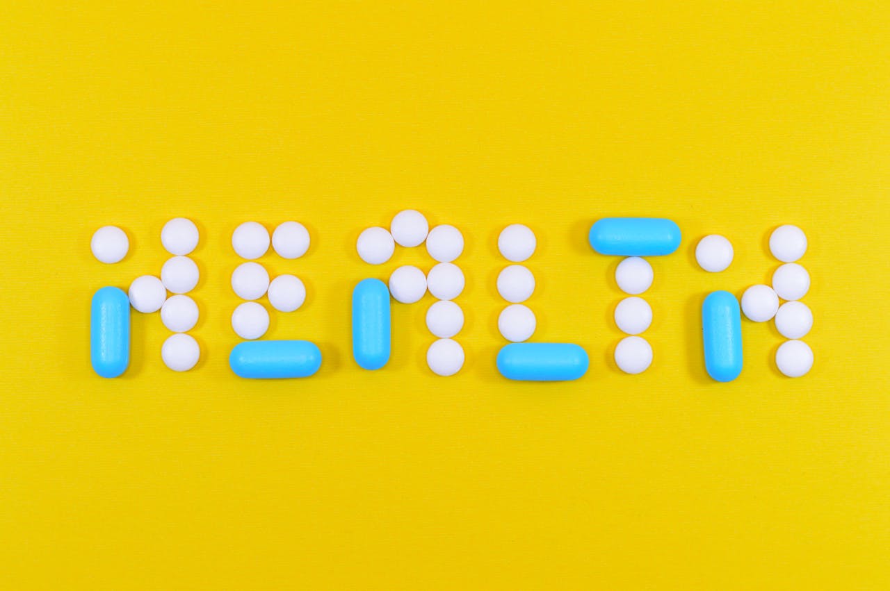 The word 'HEALTH' spelled out with white round pills and blue capsule pills on a yellow background.
