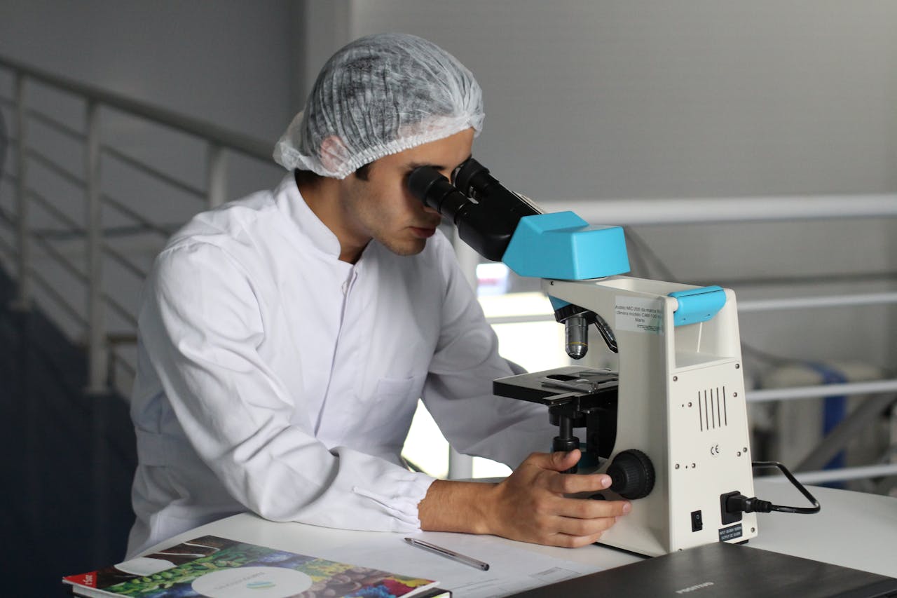 Scientist in a white lab coat and hairnet looking through a microscope in a laboratory setting.