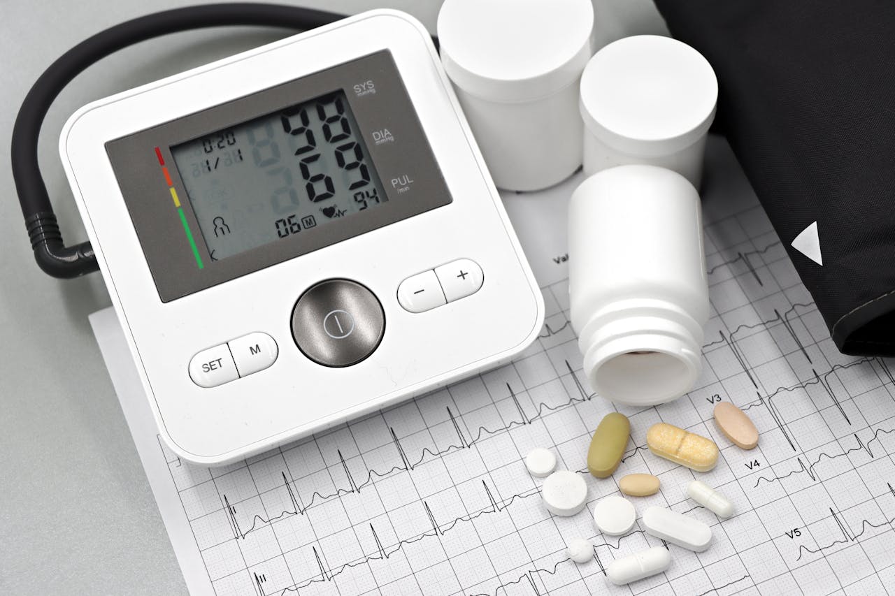 Digital blood pressure monitor showing 98/69 with heart rate 94, surrounded by various pills on an ECG printout and a blood pressure cuff.