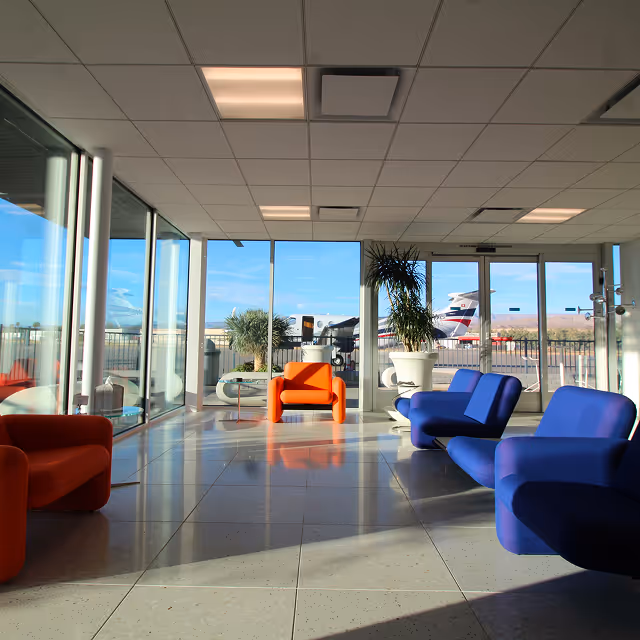 Dunes Air Modern airport lounge with glass walls, featuring orange and blue armchairs and a view of an airplane outside.