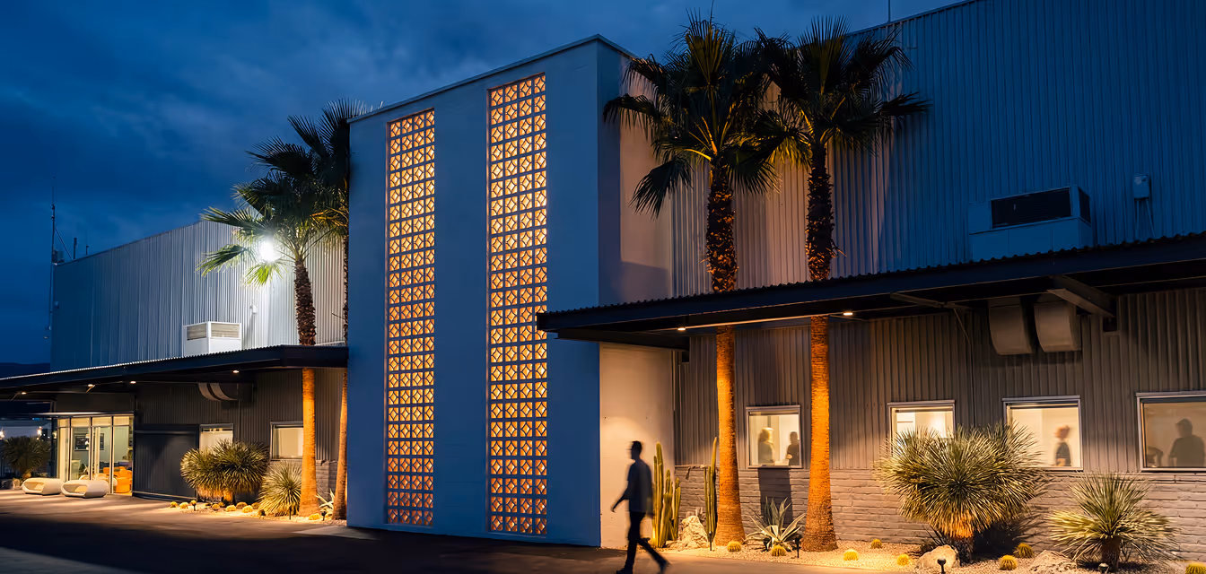 Dunes Air Modern industrial FBO exterior at dusk with illuminated decorative wall panels, palm trees, and silhouettes of people visible through windows.