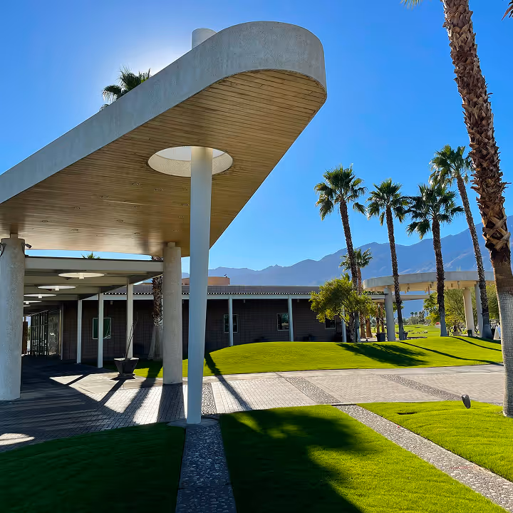 Modern building with curved concrete canopy and palm trees on a sunny day with mountains in the background.