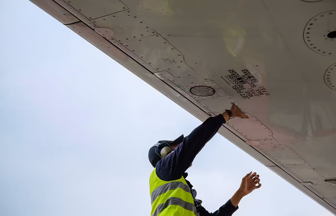 Aircraft maintenance worker in a yellow safety vest inspecting the underside of an airplane wing.
