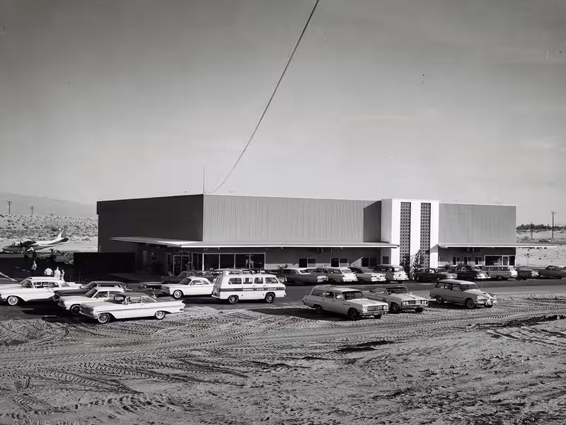 Dunes Air FBO Black and white photo mid-century modern FBO with vintage cars parked in front and an airplane visible on the left side.