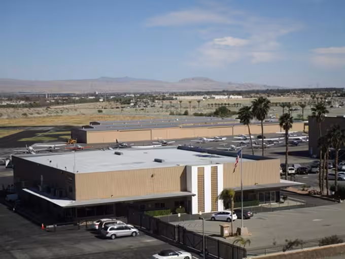 Aerial view of the Dunes Air FBO with parked cars in front, palm trees, and mountains in the background under a clear blue sky.