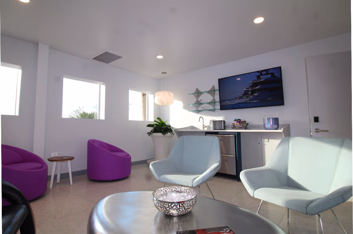 Dunes Air Modern lounge area with blue and purple armchairs, a round table with a decorative bowl, a kitchenette with appliances, and a wall-mounted TV.