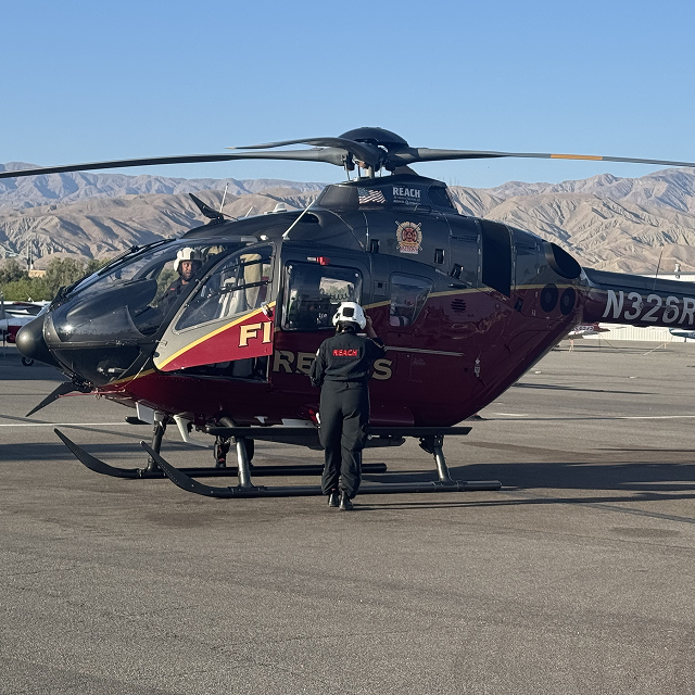 Fire rescue helicopter on tarmac with a person in a helmet standing beside it and mountains in the background.