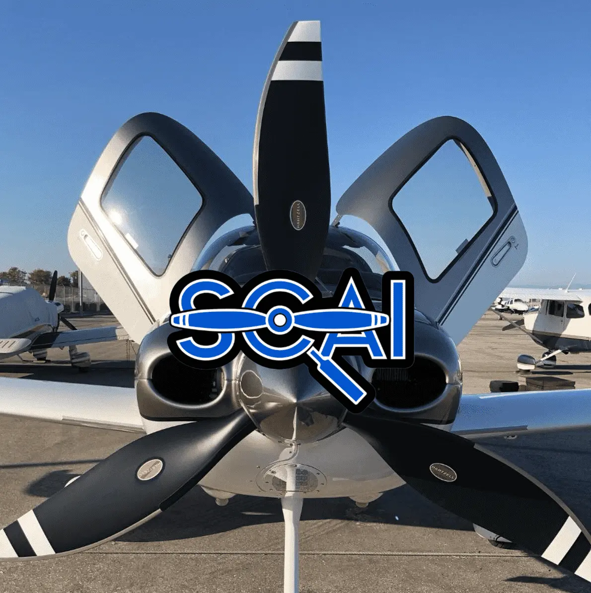 SOCAL Maintenance, front view of a small airplane with open cockpit doors and three-blade propeller on an airport tarmac under a clear sky.