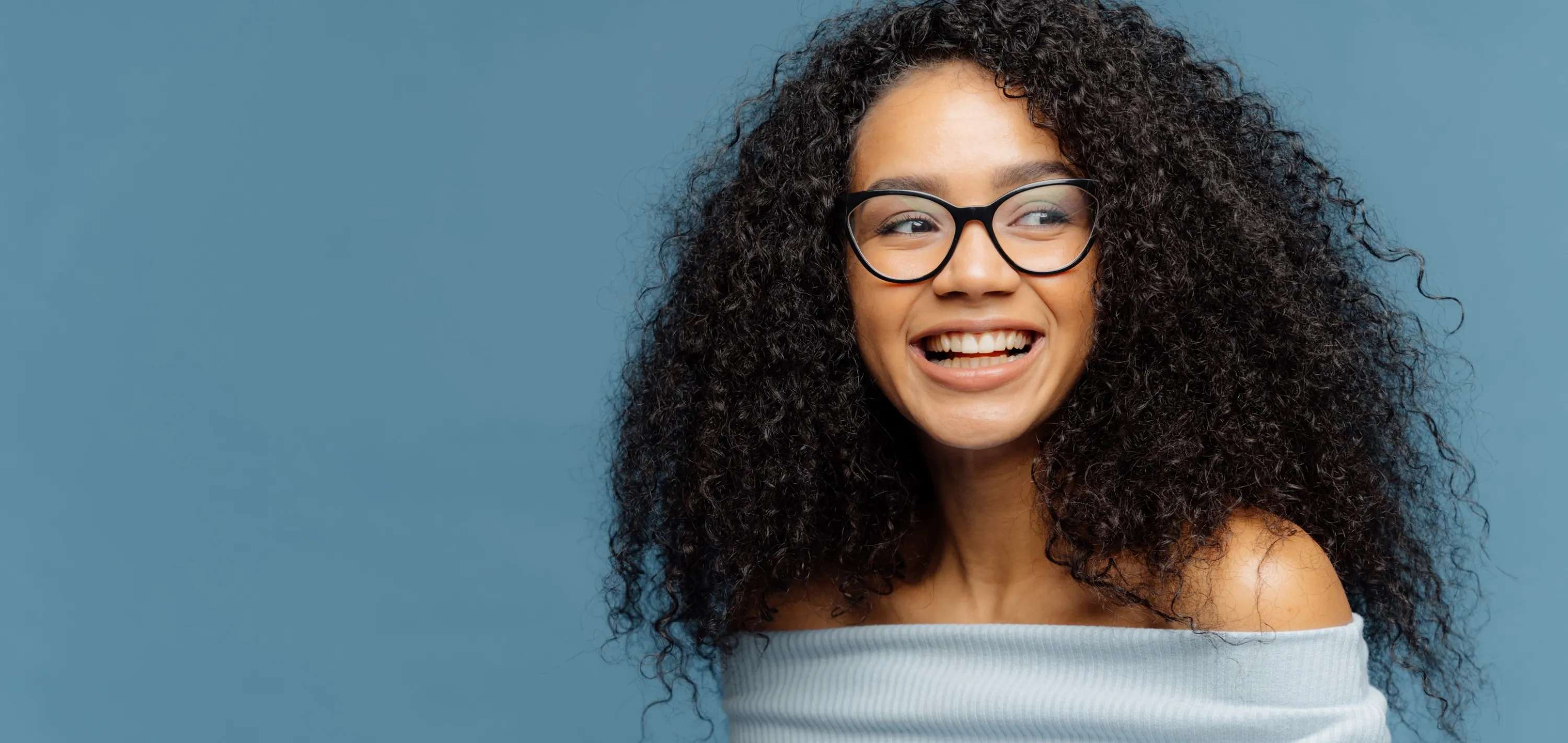 Smiling woman with curly hair, black glasses, and an off-shoulder white top against a blue background.