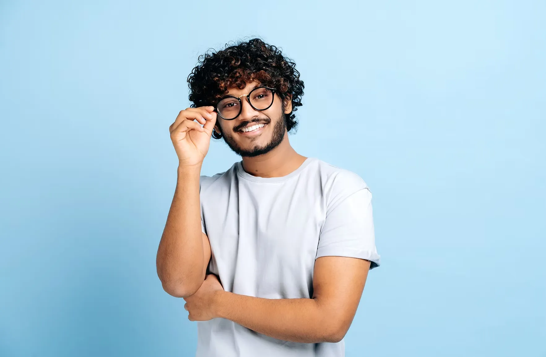 Smiling man with curly hair and glasses adjusting his frame against a light blue background.