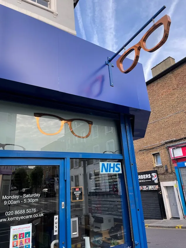 Blue storefront with large orange eyeglasses sign above and on window; NHS sticker on the door.