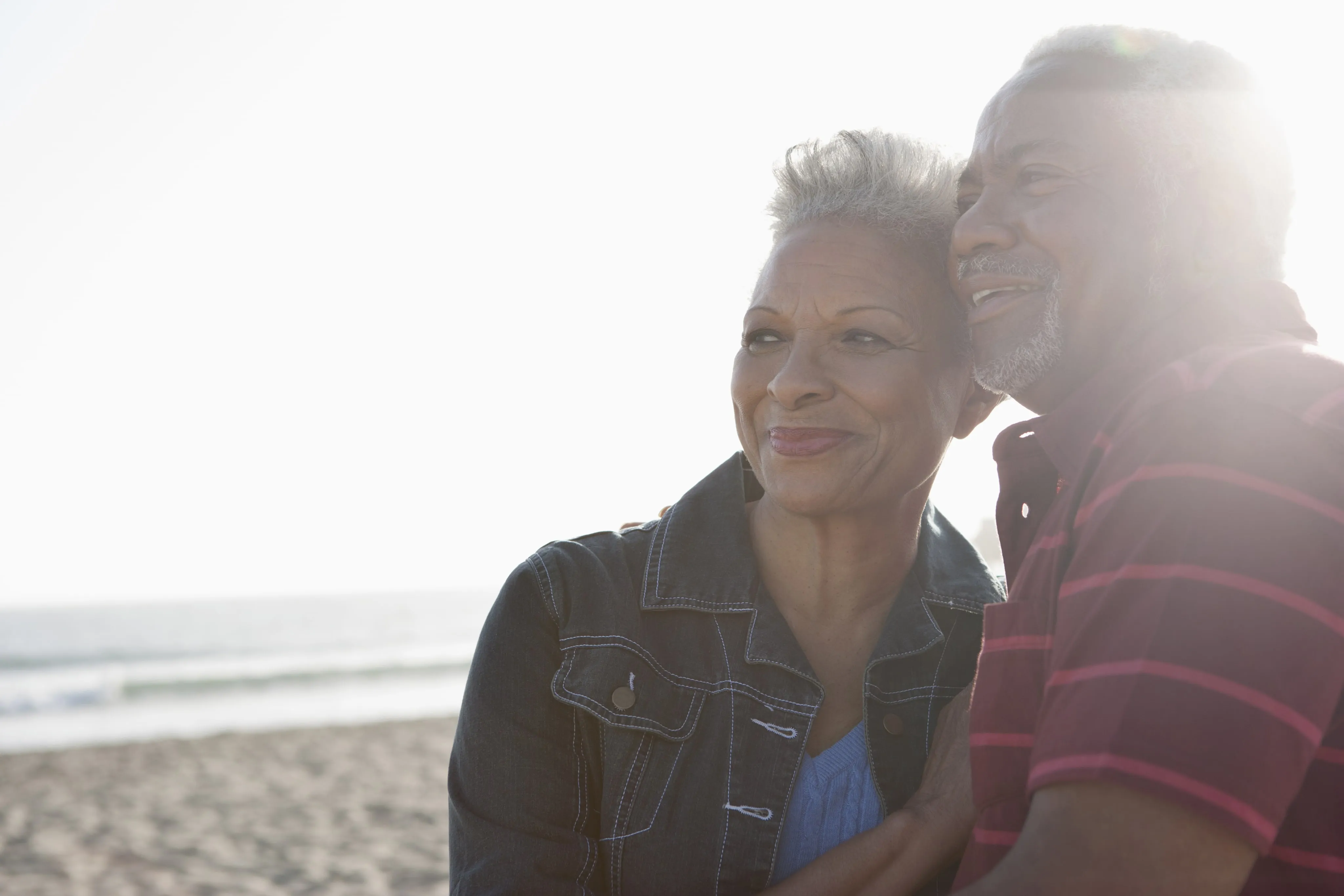Older couple smiling and embracing on a beach with the ocean in the background.