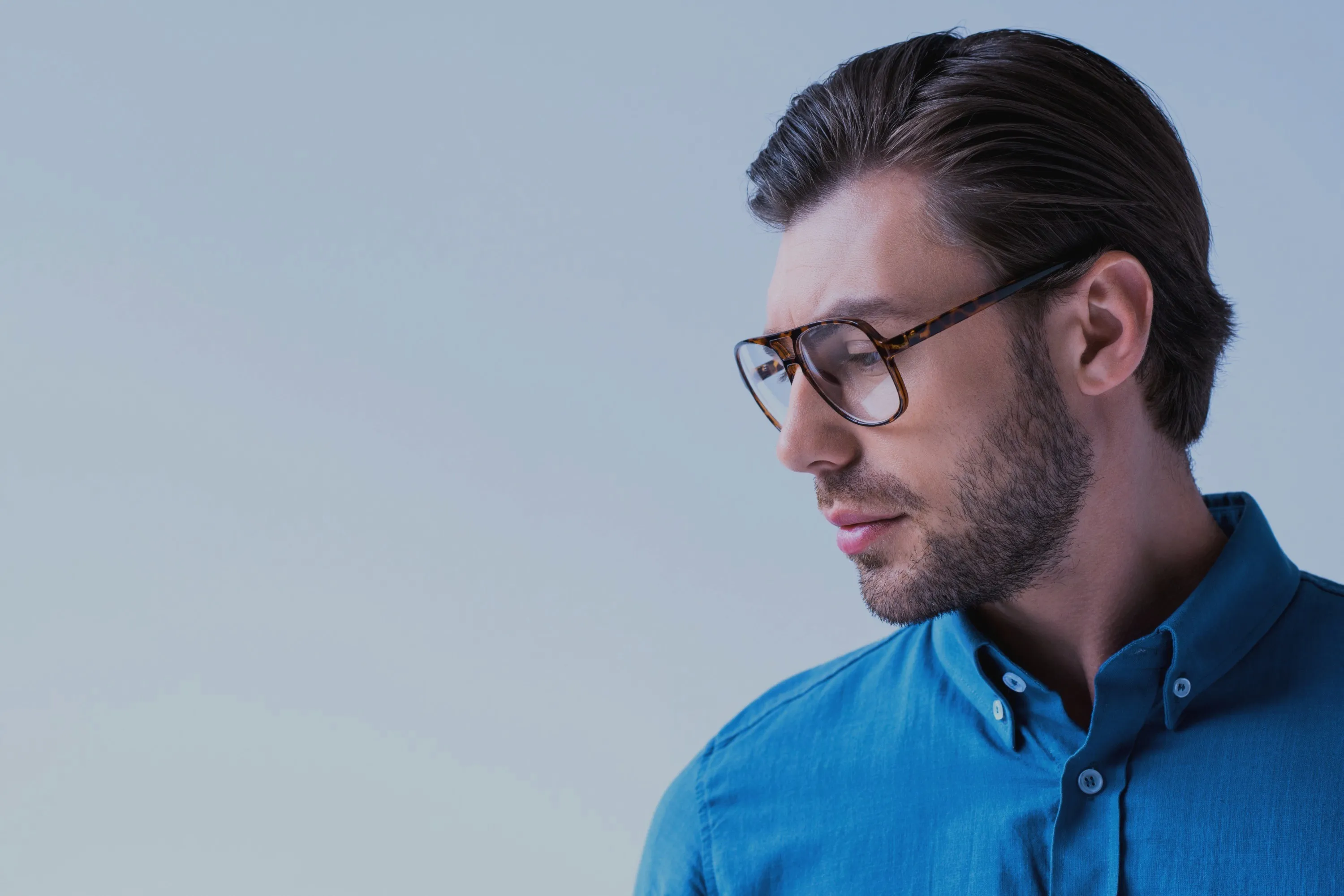 Profile of a man wearing tortoiseshell glasses and a blue button-up shirt looking downwards.