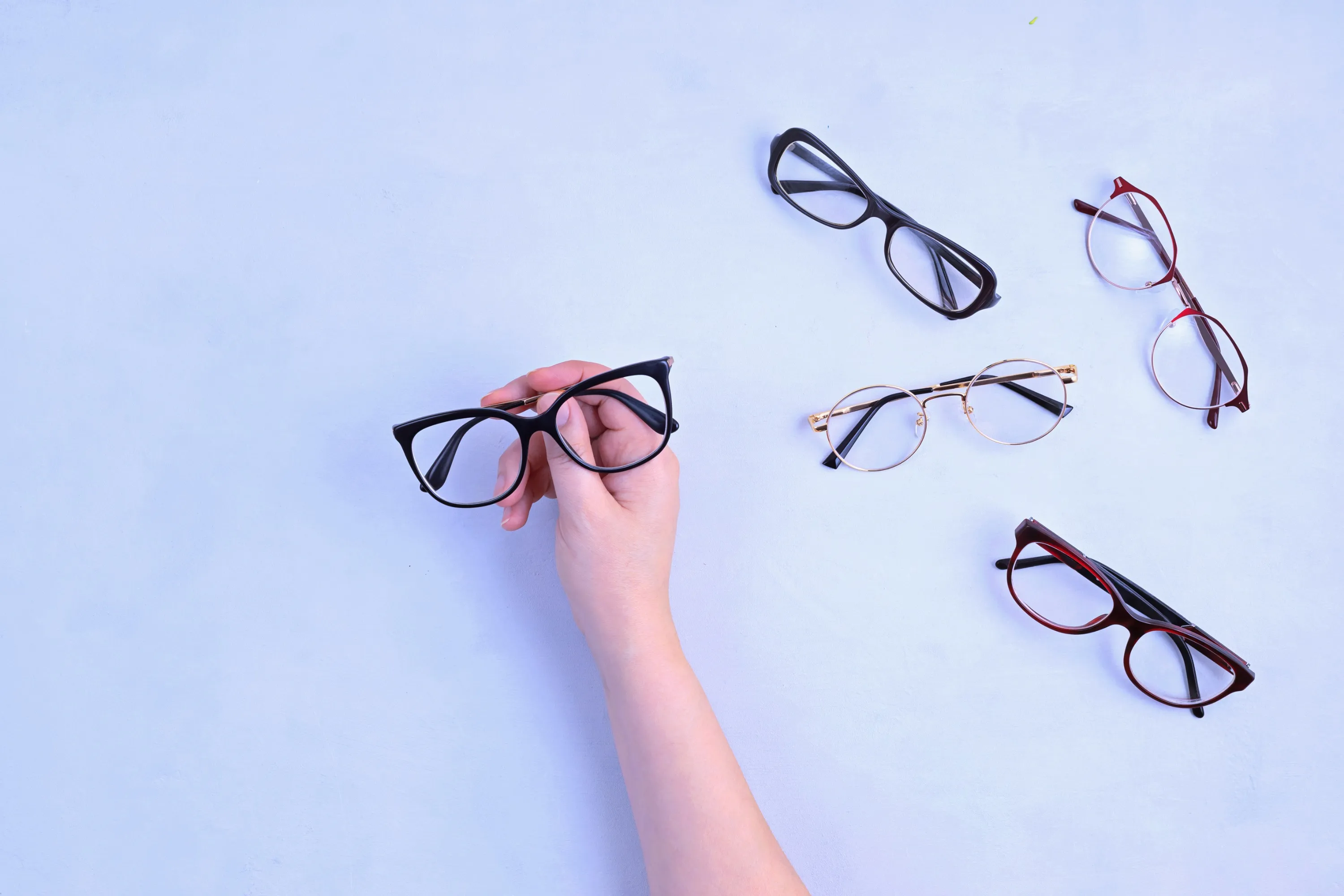 Hand holding a pair of black-framed eyeglasses with four other pairs of eyeglasses on a light surface.