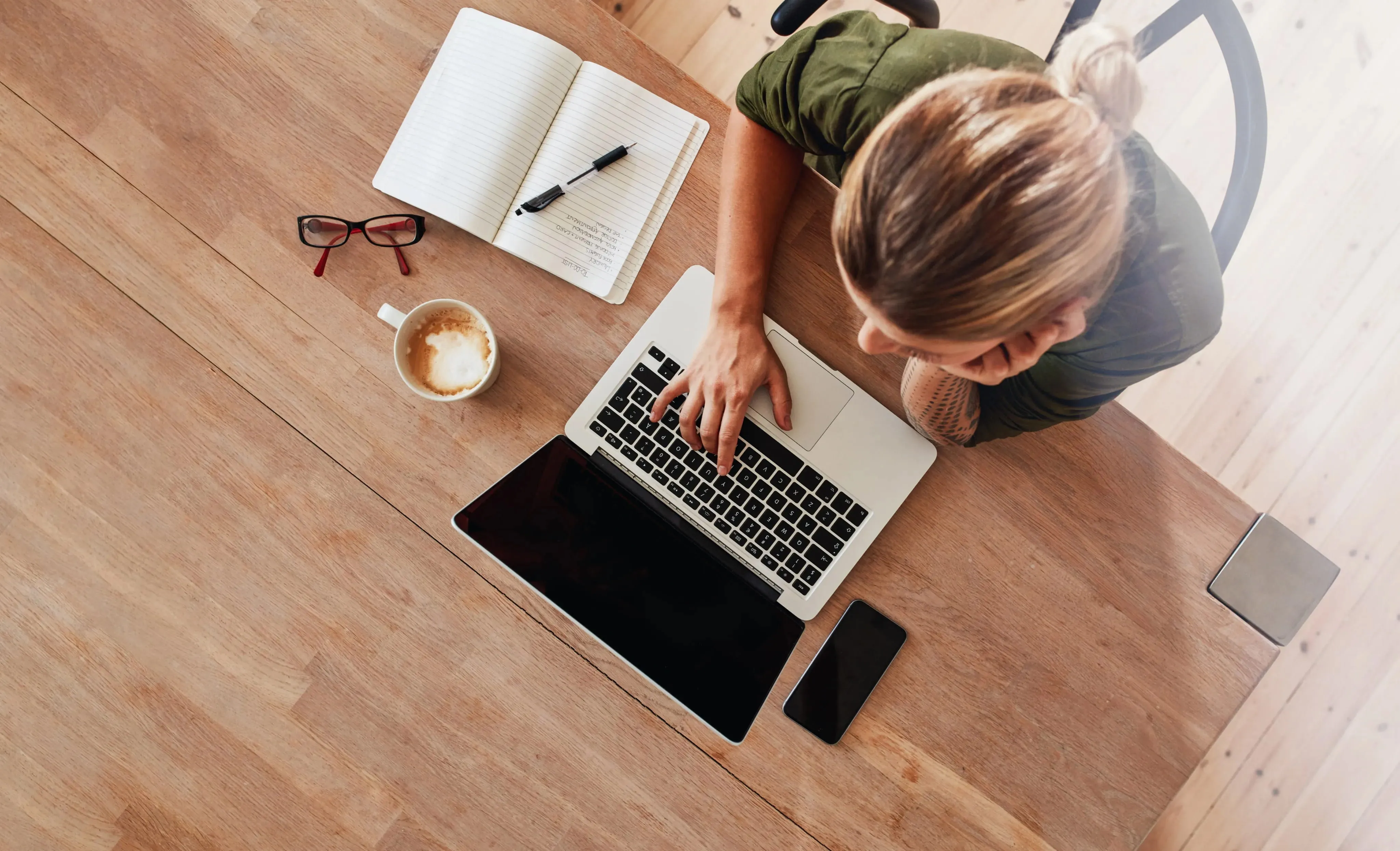 Overhead view of a person with blonde hair and a tattoo, working on a laptop at a wooden table with a notebook, glasses, coffee cup, and smartphone.