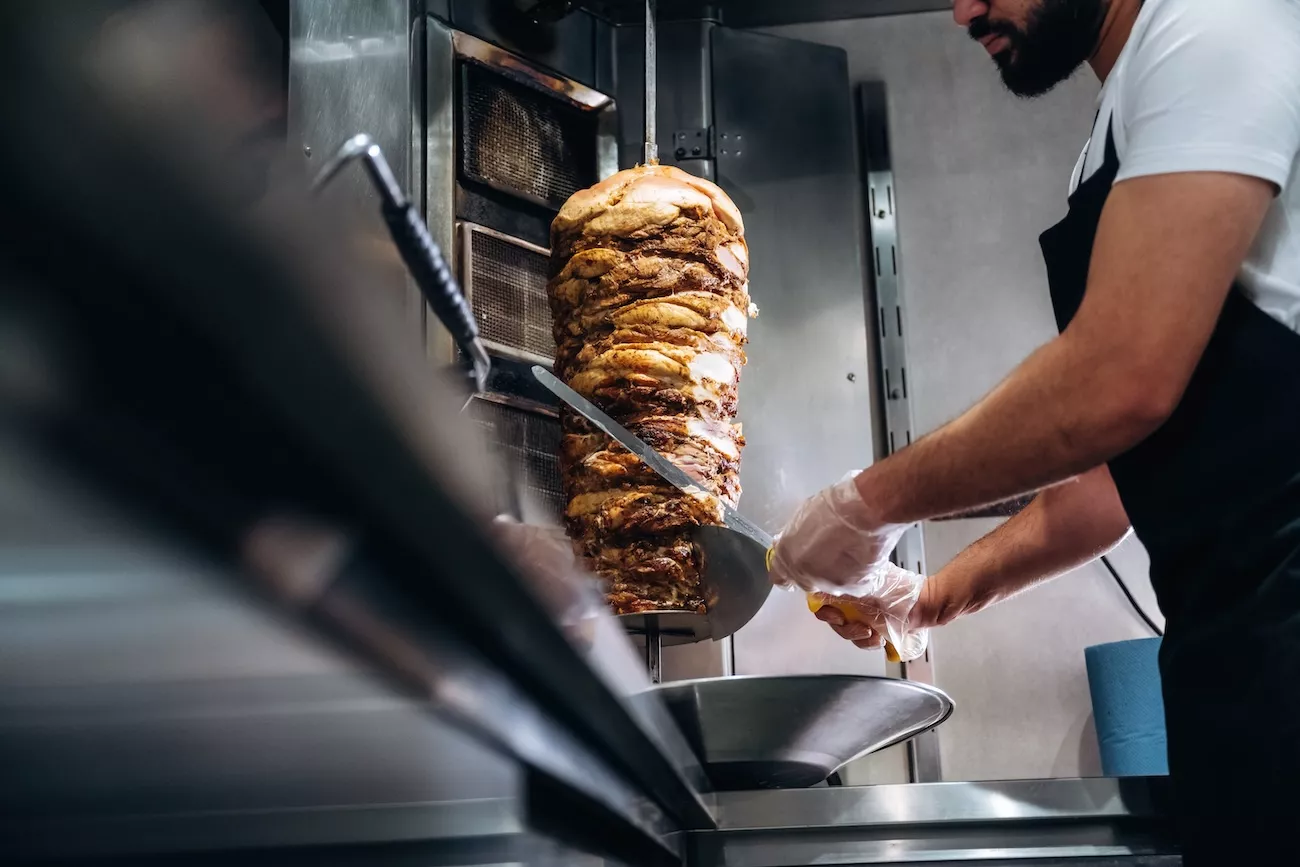 Chef slicing doner kebab meat from a vertical rotisserie grill in a commercial kebab shop kitchen