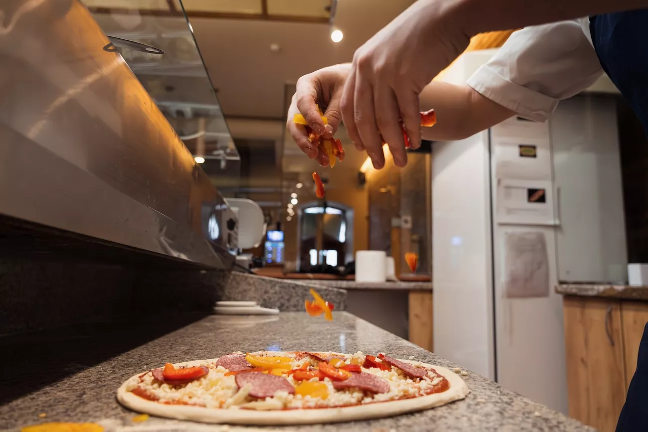 Chef adding fresh pepper and salami toppings to a pizza base in a commercial pizza shop kitchen.