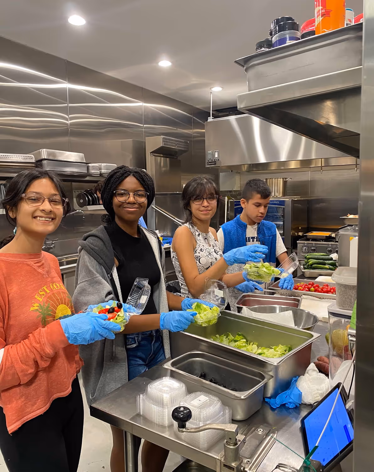 Four young people wearing blue gloves packing salad into plastic containers in a commercial kitchen.