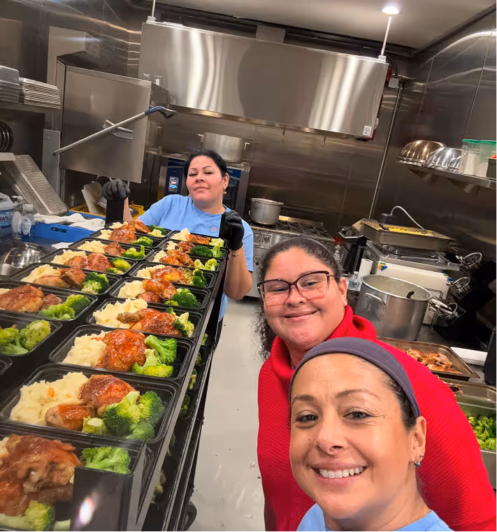 Three women smiling in a kitchen with trays of meals containing roasted chicken, mashed potatoes, and broccoli.