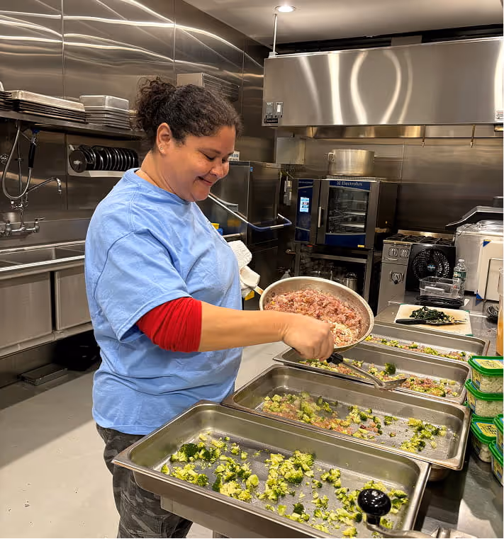 Smiling woman in blue shirt and red sleeve spreads a mixture onto trays of broccoli in a stainless steel kitchen.