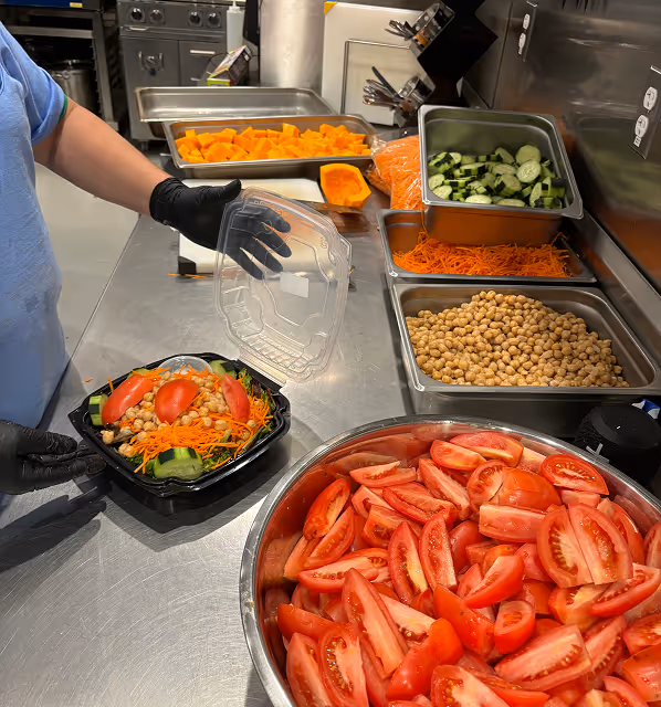 Person with black gloves packing a salad with chickpeas, tomatoes, shredded carrots, and cucumbers in a black container on a stainless steel kitchen counter, with trays of pre-cut vegetables nearby.
