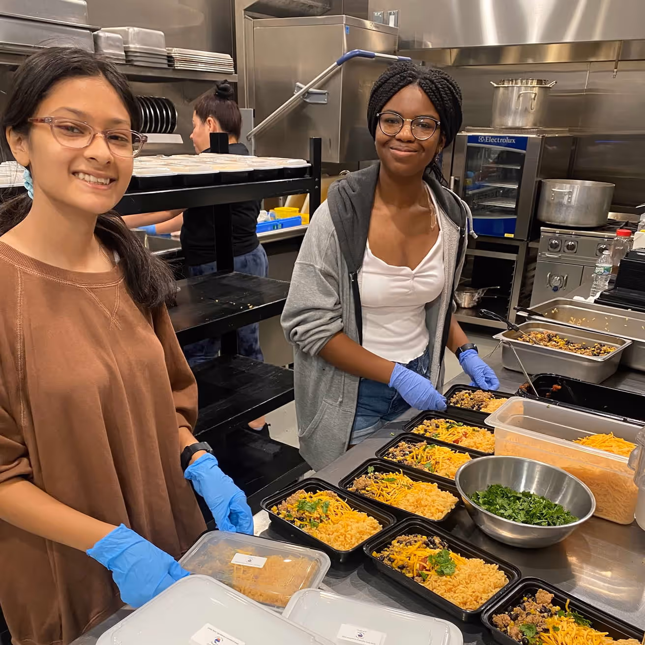 Two young women wearing gloves smiling and preparing multiple meal containers with rice, beans, and cheese in a commercial kitchen.