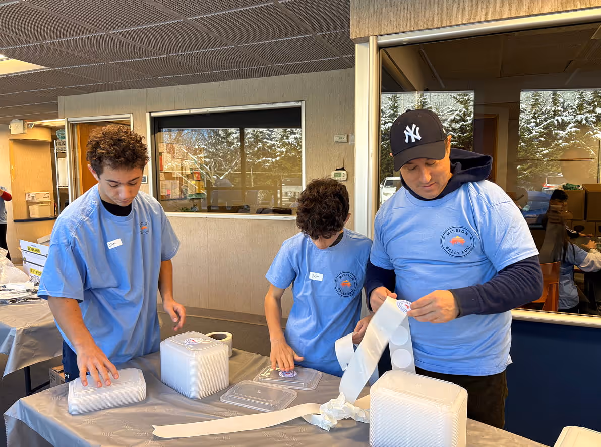 Man wearing a blue shirt working at a stainless steel sink in a commercial kitchen with food prep tables and cooking equipment.