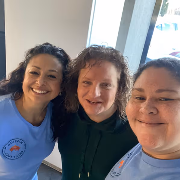 Three smiling women standing indoors near a window, two wearing light blue shirts with logos and the woman in the middle wearing a dark green hoodie.