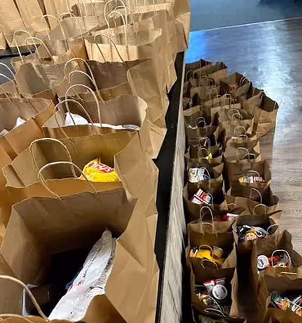 Rows of brown paper bags filled with assorted groceries and snacks on a floor and table.