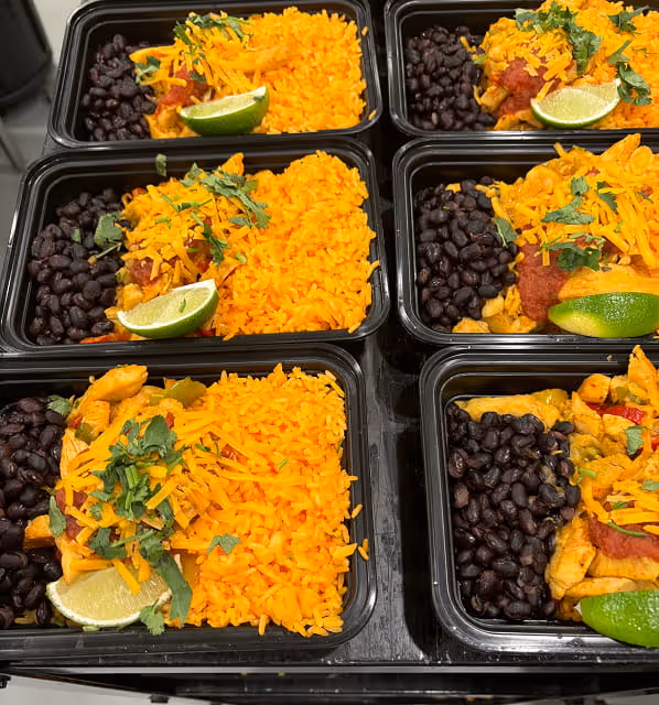 Meal prep containers filled with yellow rice, black beans, seasoned chicken topped with shredded cheese and herbs, and lime wedges.