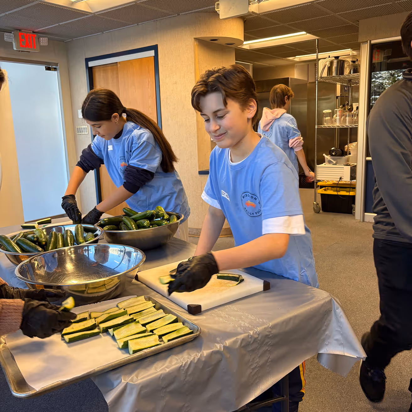 Two young volunteers in blue shirts and black gloves slicing zucchinis in a kitchen setting.