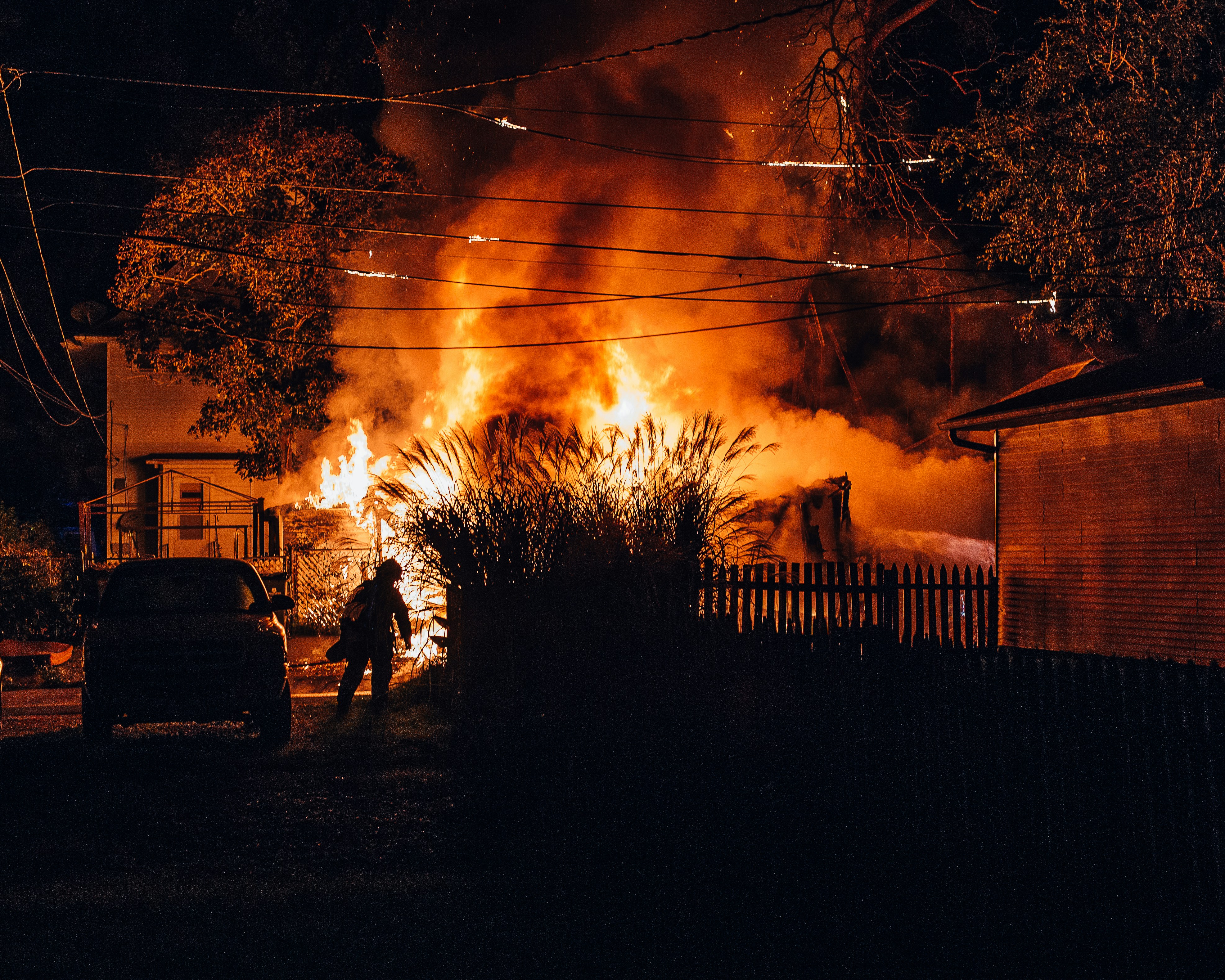 Large house engulfed in flames at night with a person silhouetted walking near the burning building.