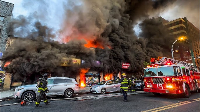 Firefighters in gear responding to a building fire with thick black smoke and flames engulfing storefronts, a fire truck on the street, and damaged cars nearby.