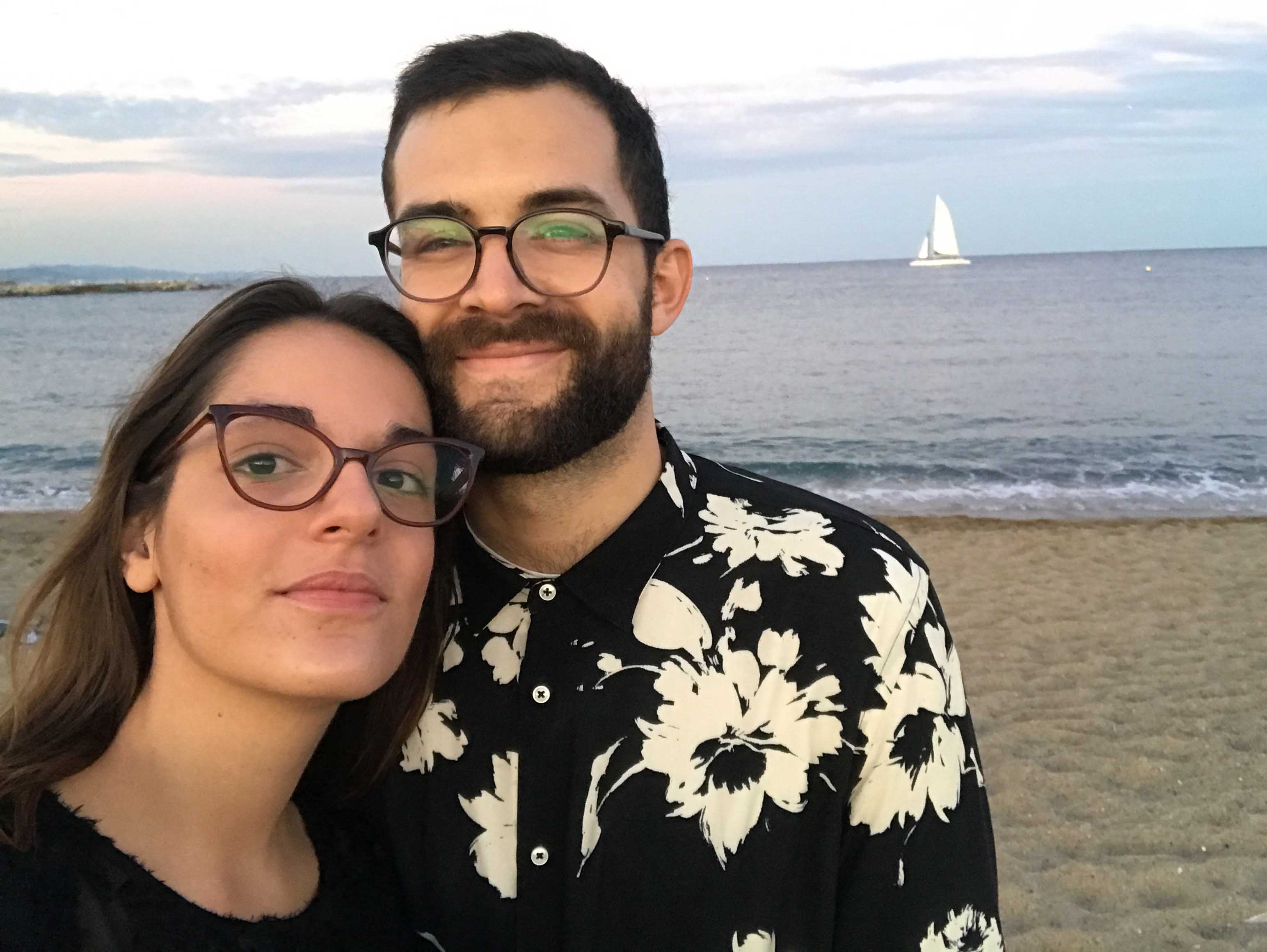 Smiling couple wearing glasses posing for a selfie on a sandy beach with the ocean and a sailboat in the background.