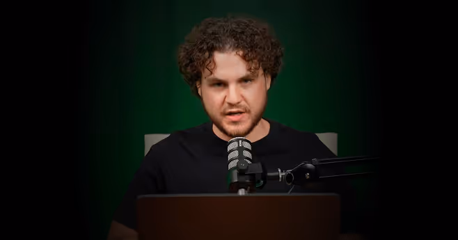 Man with curly hair speaking into a microphone behind a laptop against a dark green background.