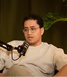 Man wearing glasses and a beige sweater speaking into a microphone with a green plant in the background.