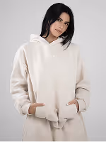 Woman with dark hair wearing an oversized cream hoodie and matching pants with hands in pockets.